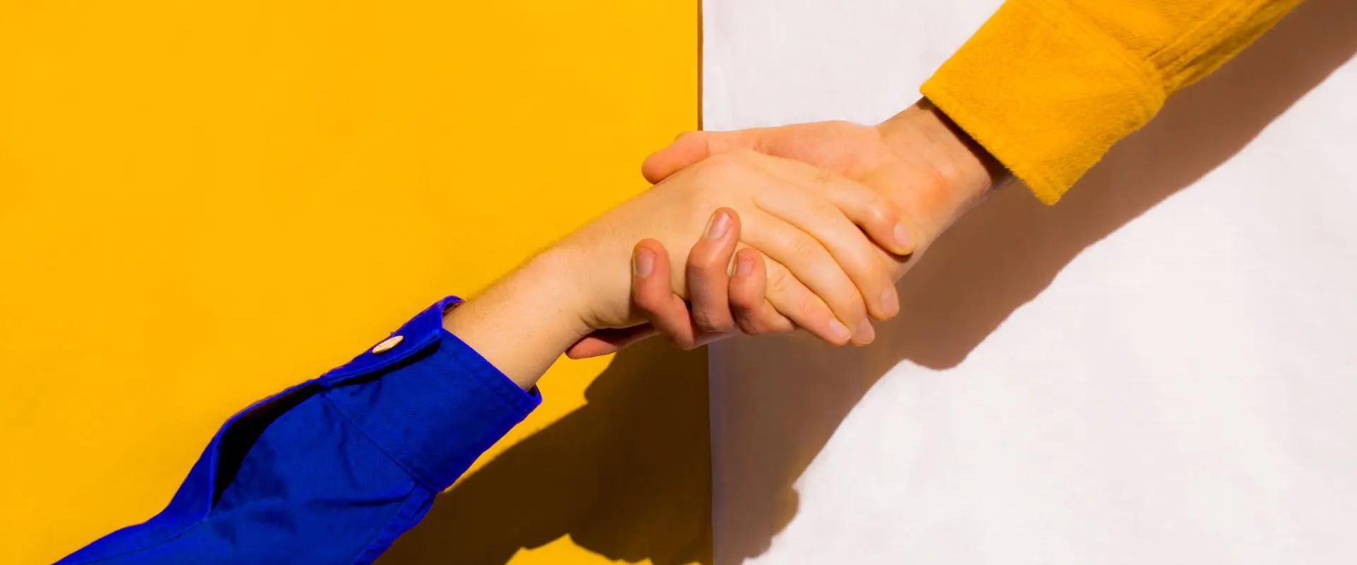 In the image two hands are shaking. The person on the right is wearing a yellow shirt and the person on the left is wearing a blue shirt.