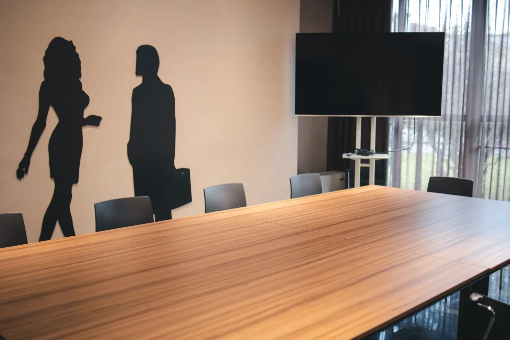 The image shows the Borghetto Room of the hotel with a large table in the center surrounded by black chairs. Behind it is a television and windows covered by curtains. On the left wall are black stickers depicting the silhouettes of two people.