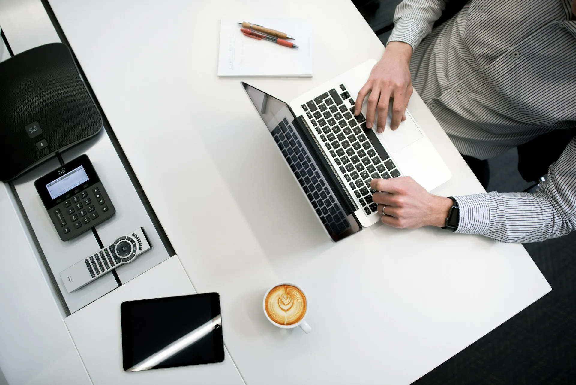 In the image a man is sitting at a desk and working on a laptop. He is wearing a white and gray checked shirt. To the right of the laptop is a notepad and two pens. On the desk is also a cappuccino, a tablet, a remote control, a keypad and a speaker.