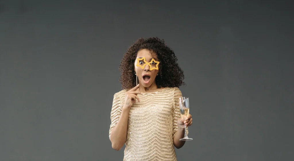 Nell'immagine una ragazza ha la bocca spalancata e la faccia sorpresa. In una mano regge un calice di vino bianco e nell'altra il bastoncino di un paio di occhiali di carta a forma di stella gialli. La ragazza indossa un abito dorato con paillettes e ha folti capelli ricci.